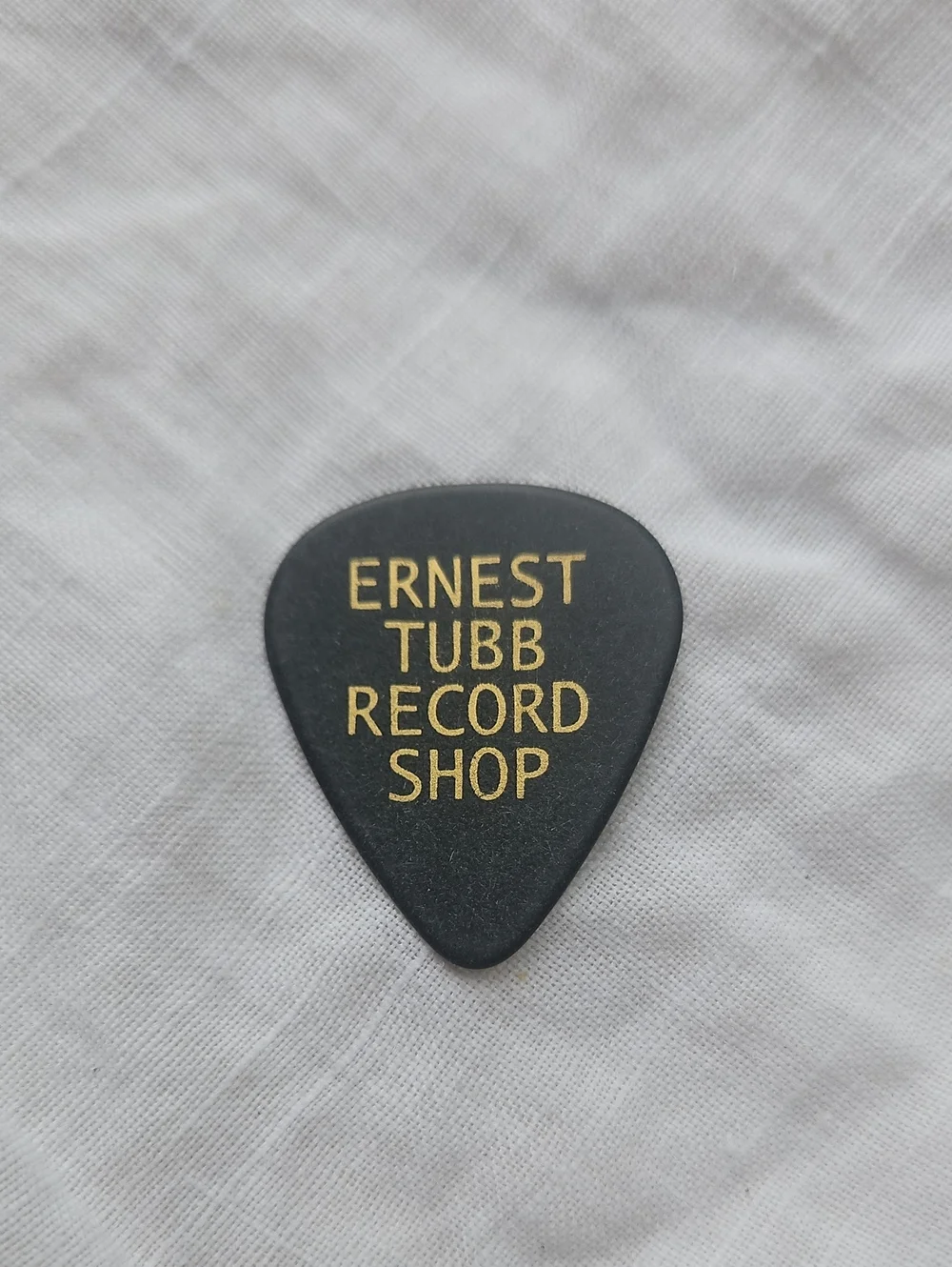 Promotional guitar pick featuring gold lettering that reads 